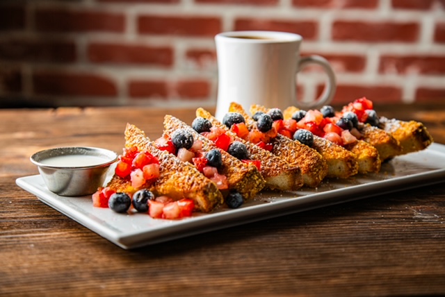 Blu Jam Cafe's signature Crunchy French Toast topped with fresh strawberries and blueberries, served with a side of cream sauce and a cup of coffee on a wooden table